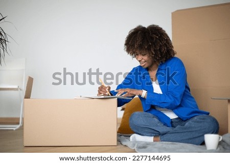 Smiling Black Female Moving Home, Writing Checklist Of Necessities For New House, Happy Young African American Woman Sitting Among Cardboard Boxes And Taking Notes To Notepad, Copy Space [[stock_photo]] © 