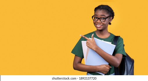 Smiling Black Female Student Pointing Finger Aside At Empty Space For Text Posing With Books And Notebooks Standing Over Yellow Studio Background. Great Educational Offer Advertisement