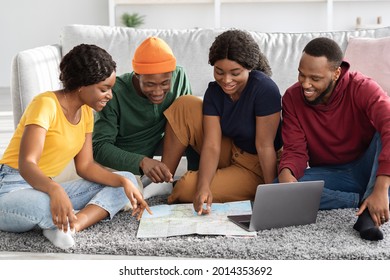 Smiling Black Friends Sitting On Floor At Home, Planning Summer Vacation, Using Map And Laptop. Group Of African American Young Men And Women Travellers Booking Flights And Hotels