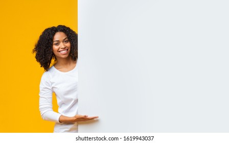 Smiling Black Girl Pointing At White Advertisement Board For Your Text With Hand, Demonstrating Copy Space, Standing Over Yellow Background