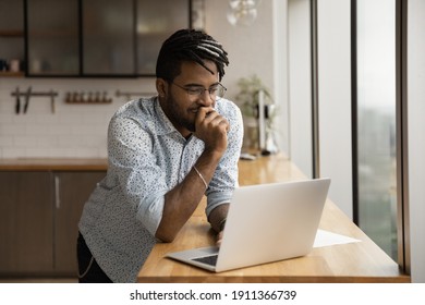 Smiling black guy freelancer stand at kitchen desk read good email message on laptop screen think on advantages proposed by customer. Young afro man hipster chat with friends online from home using pc