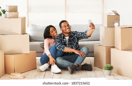 Smiling Black Guy And His Girlfriend Moving To Their Own Home Together, Taking Selfie Among Carton Boxes, Panorama. Happy African American Couple Making Photo Of First Day In New Apartment