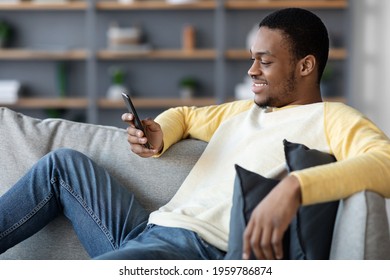 Smiling Black Guy Reclining On Couch At Home, Using Smartphone, Chatting With Friends Or Playing Online, Copy Space. Happy African American Young Man Websurfing Or Using Social Media On Mobile Phone