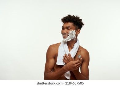 Smiling black guy with shaving foam on face holding razor and looking away. Young man with towel and naked torso. Hygiene and skin care. Isolated on white background. Studio shoot. Copy space