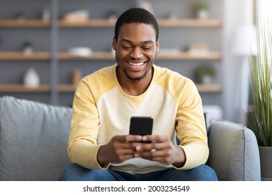 Smiling Black Guy Sitting On Couch At Home, Using Smartphone, Chatting With Friends Or Playing Online, Copy Space. Happy African American Young Man Websurfing Or Using Social Media On Mobile Phone