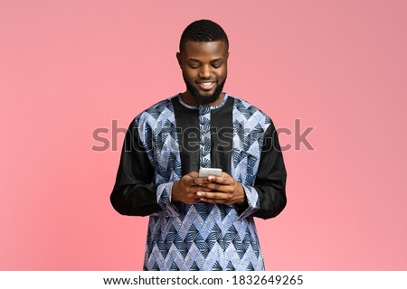 Smiling black guy using modern smartphone, wearing african t-shirt, pink studio background. African american young man in traditional costume chatting with friends on mobile phone, copy space