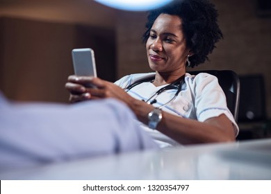 Smiling Black Healthcare Worker Enjoying In A Break And Text Messaging On Smart Phone. 