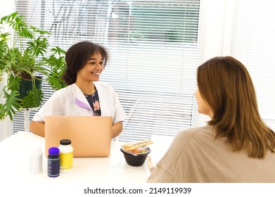 Smiling Black Lady Doctor Chatting With Female Patient At Clinic Advicing Her Vitamins And Fresh Takeaway Salad In A Lunch Box. Dietology And Nutririon Concept