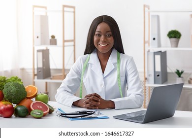 Smiling Black Lady Doctor Nutritionist Sitting At Workplace With Vegetable And Fruit, Using Laptop In Office