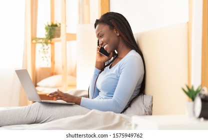 Smiling Black Lady Having Phone Conversation Using Laptop Sitting In Bed At Home. Selective Focus