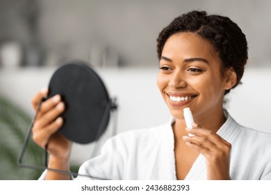 Smiling Black Lady Holding Mirror And Applying Moisturising Lip Balm, Beautiful African American Female Using Chapstick, Putting On Hygienical Lipstick While Standing In Bathroom At Home - Powered by Shutterstock