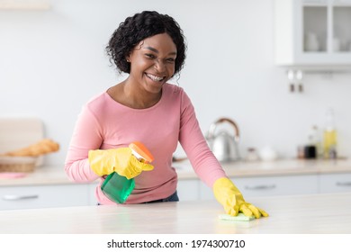 Smiling black lady housewife cleaning kitchen furniture, using spray bottle with disinfestor and dust cloth, wearing colorful rubber gloves, copy space. Young african american woman cleaning house
