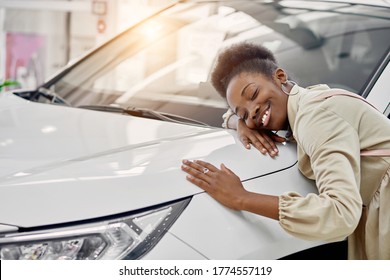 Smiling Black Lady Hugs Her New Auto In Cars Showroom. Young Woman Fulfill Her Dream, Lies On Car