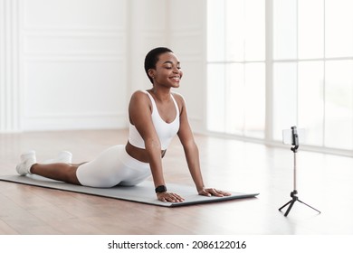 Smiling Black Lady Practicing Pilates Using Cell Phone On Tripod, Recording Video Or Watching Online Tutorials, Excercising In Living Room, Stretching Back Muscles, Standing In Upward-Facing Dog Pose