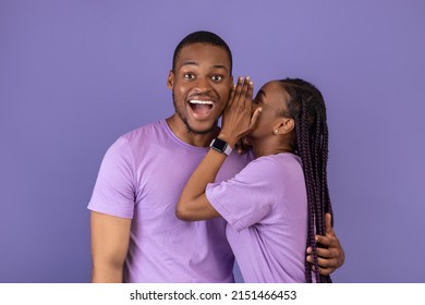 Smiling black lady sharing secret or whispering gossips into her boyfriend's ear on violet purple studio background. Millennial African American couple sharing shocking news, discussing rumors