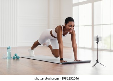 Smiling Black Lady In White Sportswear Training At Home In Front Of Cellphone On Tripod, Beautiful Female Fitness Blogger Shooting Tutorials, Doing Cross Body Mountain Climbers Exercise At Camera