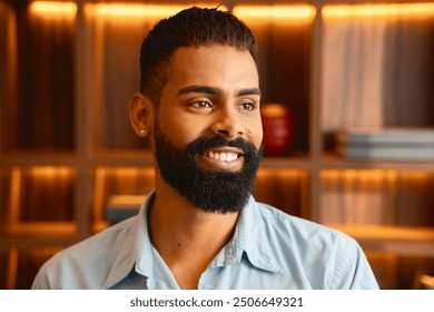 Smiling black man with a beard posing indoors, wearing a light blue shirt. Cheerful young male with stylish haircut and piercing, standing against a warm, wooden background - Powered by Shutterstock