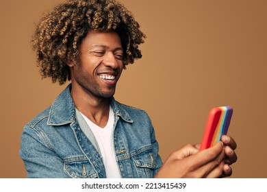 Smiling Black Man With Curly Hair And In Denim Jacket Browsing Mobile Phone In Trendy Rainbow Colored Case Supporting LGBTQ Community