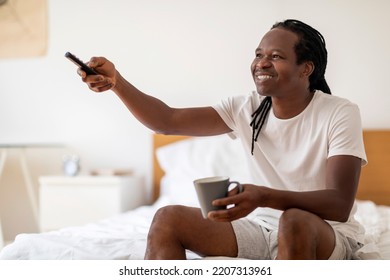 Smiling Black Man Drinking Coffee And Watching TV While Relaxing At Home, Happy African American Guy Sitting On Bed In Bedroom And Switching Channels With Remote Controller, Enjoying Domestic Pastime