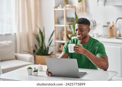 Smiling black man enjoying cup of coffee while using laptop in kitchen, freelancer at home - Powered by Shutterstock