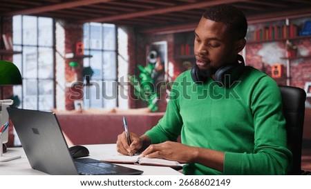 Smiling black man taking notes while watching a tutorial presentation, doing online self learning and writing details. Young adult freelancer writes in his textbook notepad. Camera B.
