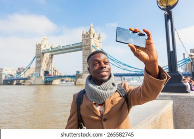 Smiling Black Man Taking Selfie In London With Tower Bridge On Background. He Is Holding The Phone And Looking At Camera. Photo Taken On A Sunny Winter Day.