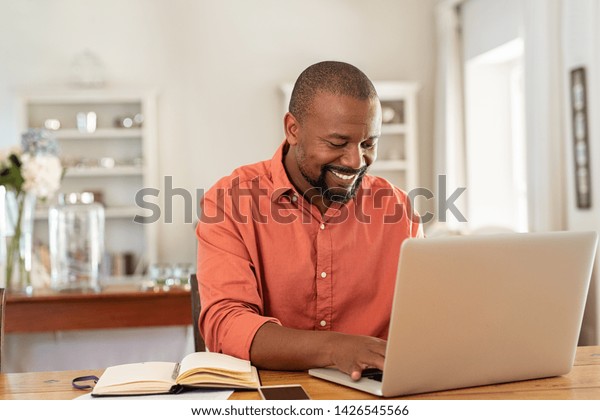 笑顔の黒人男性が自宅のリビングでノートパソコン を使う 幸せな成熟した実業家がメールを送り 自宅で働く 机の上に書類や書類を置いたコンピューターでのアフリカ系アメリカ人のフリーランス の写真素材 今すぐ編集