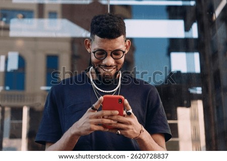Smiling Black man using red phone while texting in city, dressed in streetwear with pearls and rings, enjoying digital connection on the go, modern and youthful urban vibe