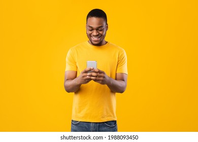 Smiling Black Man Using Smartphone Browsing Internet And Scrolling Social Media News Feed Standing Over Yellow Studio Background. Millennial Guy Using New Mobile Application. People And Gadgets