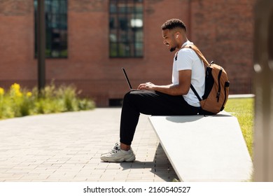 Smiling Black Man In Wireless Earphones Working On Laptop Computer Outdoors, Cheerful Guy Sitting In College Campus And Using Pc, Looking At Screen, Enjoying Freelance Remote Job Or Distance Education