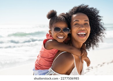 Smiling black mother and beautiful daughter wearing sunglasses having fun on the beach. Portrait of happy african american woman giving a piggyback ride to her cute little girl wearing shades.
