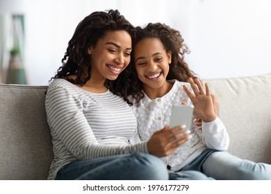 Smiling Black Mother And Daughter Having Video Call On Smartphone, Sitting On Couch And Waving At Gadget Screen. Happy African American Mom And Kid Having Online Party With Family, Copy Space