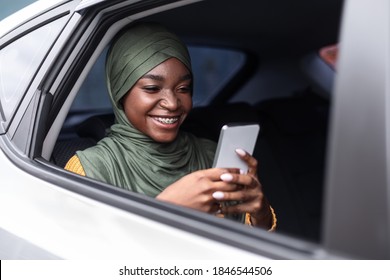 Smiling Black Muslim Lady In Hijab Using Smartphone While Riding Car On Backseat, Browsing Internet Or Texting With Friends, African Woman Enjoying Online Communication, Closeup Shot Through Window