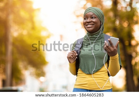 Smiling black muslim woman in headscarf walking by street and listening music on smartphone, wearing earphones and carrying backpack, positive african islamic lady enjoying spending time outdoors