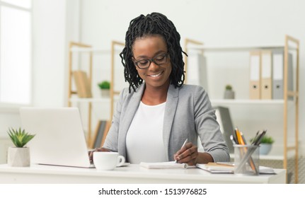 Smiling Black Office Girl Sitting At Laptop Taking Notes At Work Indoor. Business Schedule Concept. Free Space