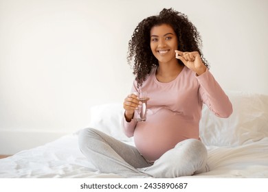 Smiling black pregnant woman showing vitamin pill and holding glass of water, african american expectant mother promoting health and prenatal care while sitting on bed in cozy bedroom at home - Powered by Shutterstock