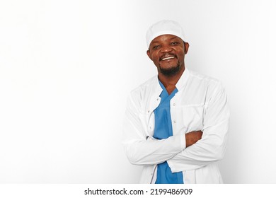 Smiling Black Surgeon Doctor Bearded Man In White Coat And Cap With Crossed Arms Isolated On White Background Copy Space. Happy Adult Black African American Practicing Surgeon Portrait Friendly Look