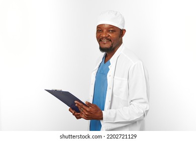 Smiling Black Surgeon Doctor Bearded Man In White Coat And Cap With Medical Records On Clipboard, Isolated On White Background Copy Space. Happy Black African American Practicing Surgeon Portrait