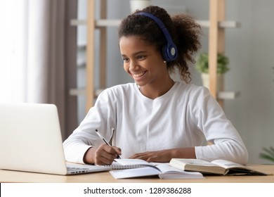 Smiling black teenage girl wearing headphones using laptop preparing homework, happy African American pupil listen to music, doing school task at home, search information online at computer