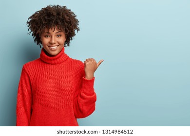 Smiling black woman with Afro hairstyle, wears red jumper, points with thumb, dressed in oversized red warm sweater, shows fantastic product, isolated over blue background, smiles from amazement