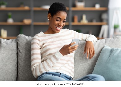 Smiling Black Woman Browsing New App On Smartphone While Resting At Home, Young Happy African American Lady Relaxing With Mobile Phone, Sitting On Couch In Cozy Living Room, Closeup Shot