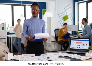 Smiling black woman entrepreneur holding clipboard with company strategy smiling at camera. Diverse team of business people analyzing company financial reports from computer.