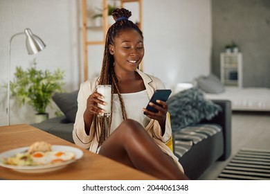 Smiling Black Woman Having A Glass Of Milk And Using Smart Phone During Her Breakfast At Home.