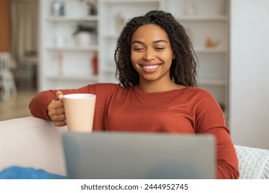 Smiling black woman sitting comfortably on a couch, holding a coffee cup, african american female focused on a laptop screen in front of her. - Powered by Shutterstock