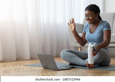 Smiling Black Woman In Sportswear Having Online Fitness Training With Laptop At Home, Waving Hand At Computer Webcam, Greeting Coach, Exercising On Yoga Mat In Living Room, Enjoying Healthy Lifestyle - Powered by Shutterstock