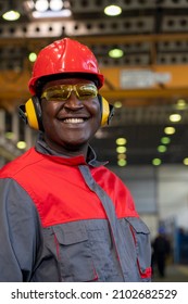 Smiling Black Worker In Personal Protective Equipment Looking At Camera. Portrait Of Industrial Worker In Red Helmet, Yellow Safety Goggles, Noise Reduction Earmuffs And Work Uniform In A Factory.