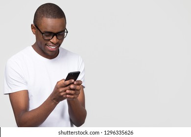 Smiling black young man in glasses holding phone looking at smartphone isolated on white grey studio background with copy space aside, happy african guy using mobile applications texting on cellphone