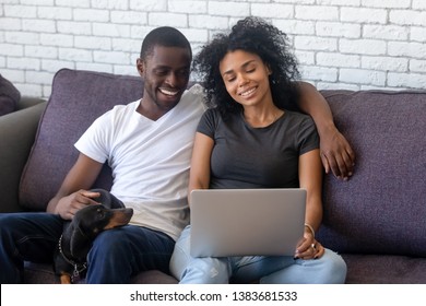 Smiling Black Young Man And Woman Relax On Couch With Dackel Dog, Hugging And Watching Movie On Laptop, Happy African American Couple Cuddle Resting On Sofa With Pet Using Computer Shopping Online