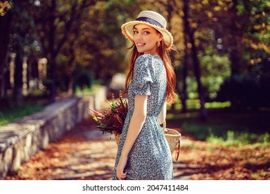Smiling blessing happy caucasian redhead girl in straw hat and fluttering dress walks alone in autumn park on sunny warm day while holds straw handbag. Indian summer concept.