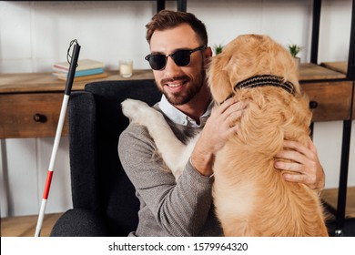 Smiling blind man sitting in armchair and hugging golden retriever at home